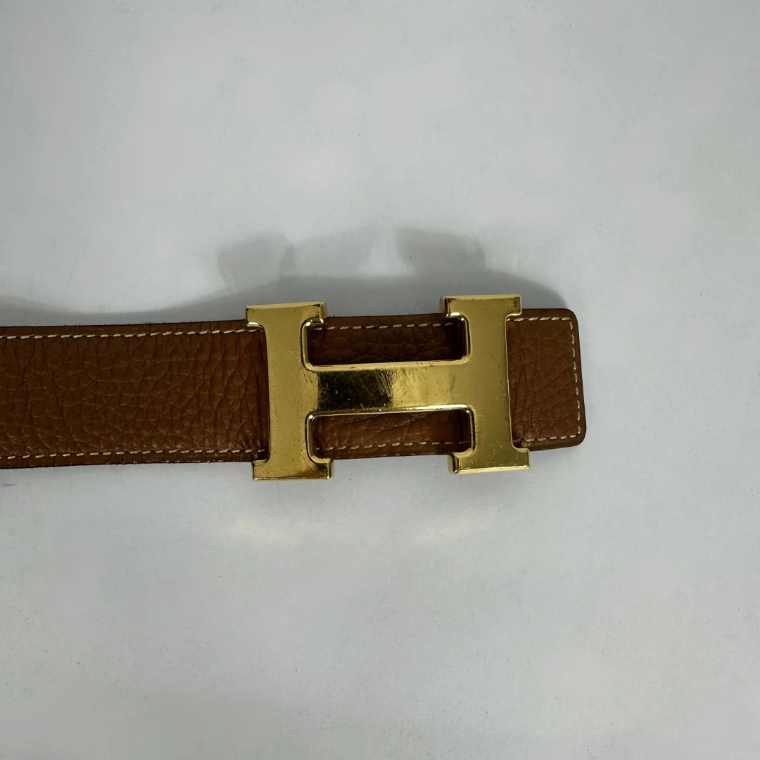 A close-up of a brown textured leather belt with a polished gold-tone H-shaped buckle, shown on a light gray background.