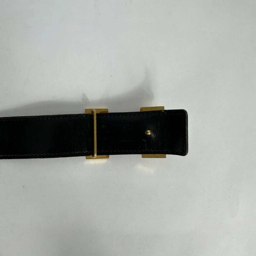 A close-up of a black leather belt with a gold-tone metal buckle, shown against a plain light gray background.