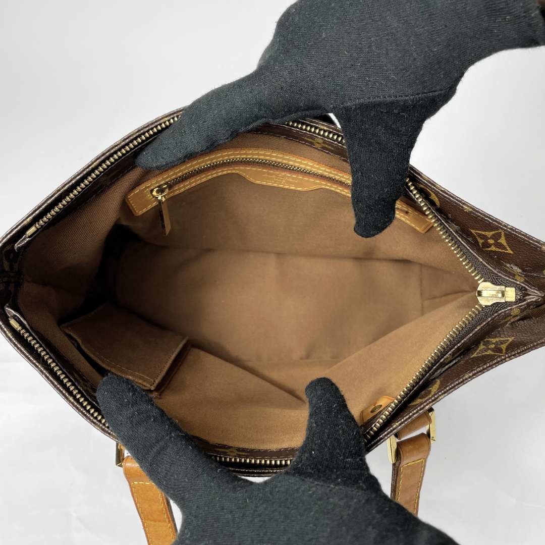 An open Louis Vuitton handbag shown from above, with black gloves holding it open to display the interior. The bag features the classic brown monogram pattern, a tan leather interior, and gold-tone zippers.