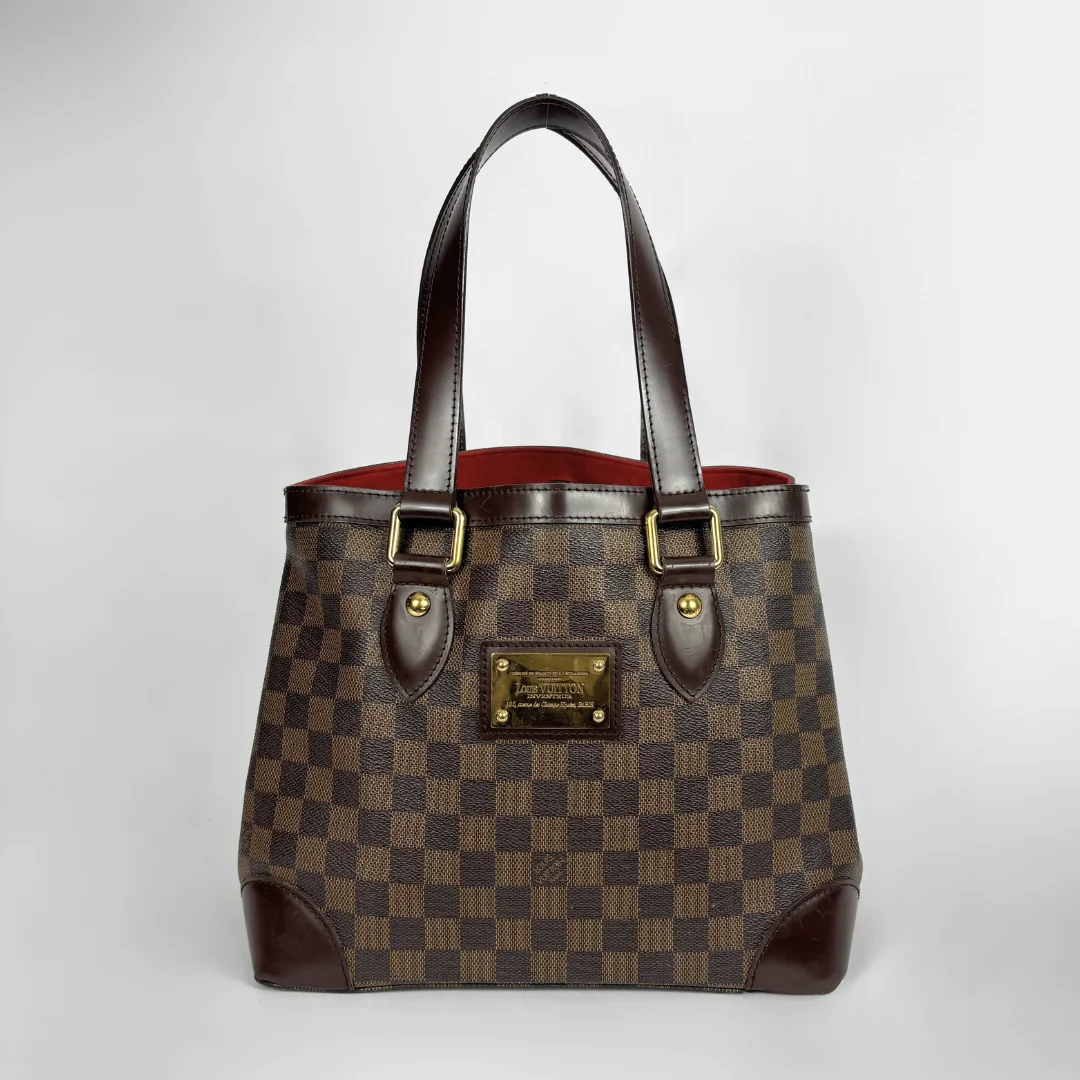 A Louis Vuitton handbag in the Damier Ebene checkered pattern, featuring dark brown leather handles and trim, gold-tone hardware, and a red interior lining, photographed against a neutral light gray background.