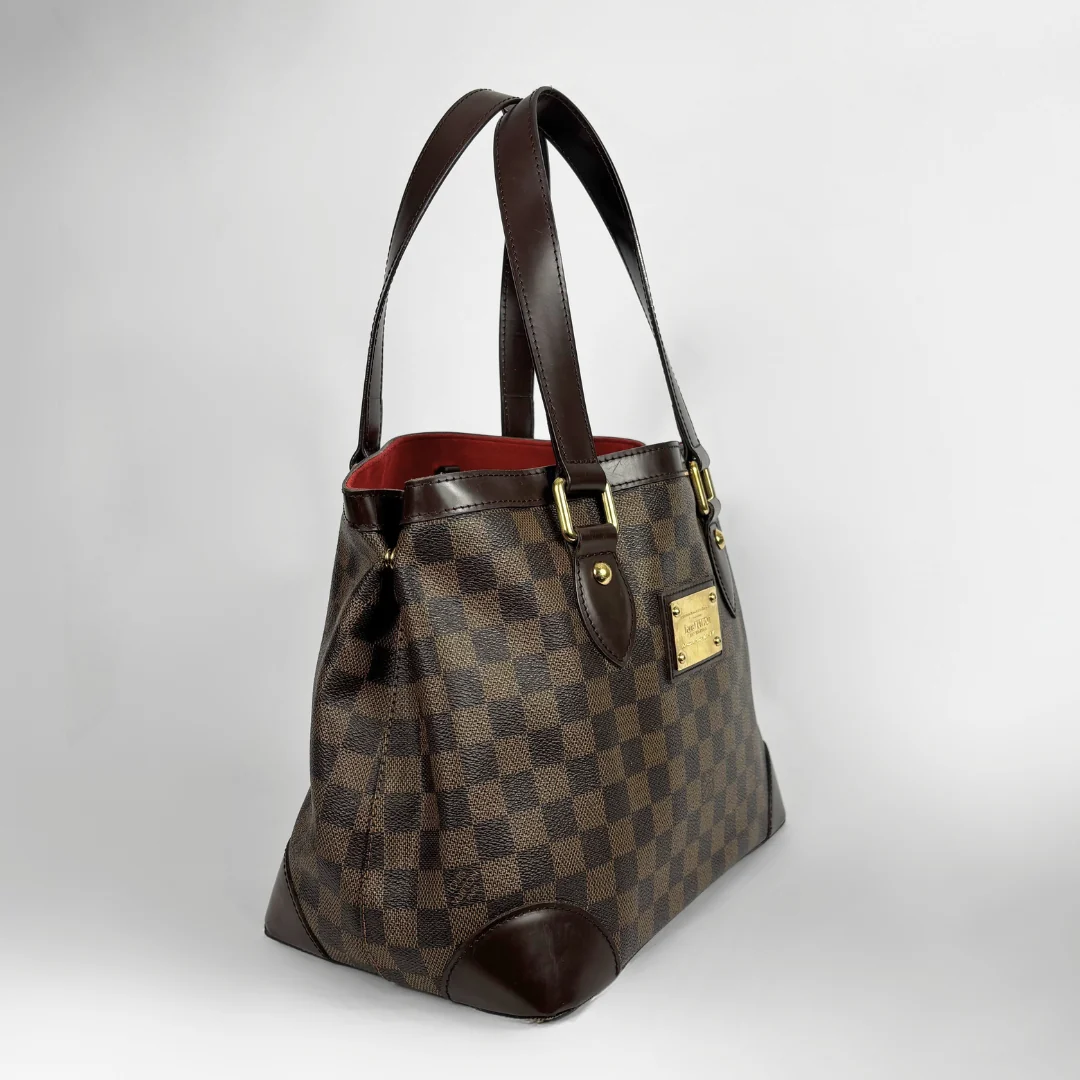 A Louis Vuitton handbag in the Damier Ebene checkered pattern, featuring dark brown leather handles and trim, gold-tone hardware, and a red interior lining, photographed against a neutral light gray background.
