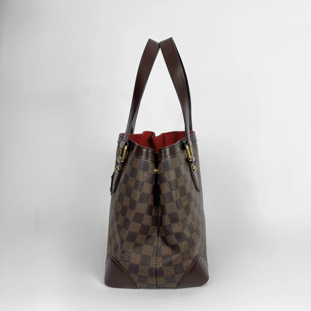 A Louis Vuitton Damier Ebene canvas tote bag with dark brown leather handles and trim, featuring a red interior lining, set against a plain light gray background.