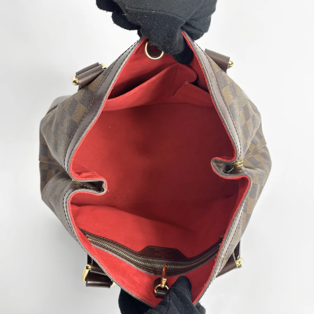 A top-down view of a Louis Vuitton handbag in Damier Ebene canvas, held open by a black glove to show the bright red interior lining and gold-tone hardware.