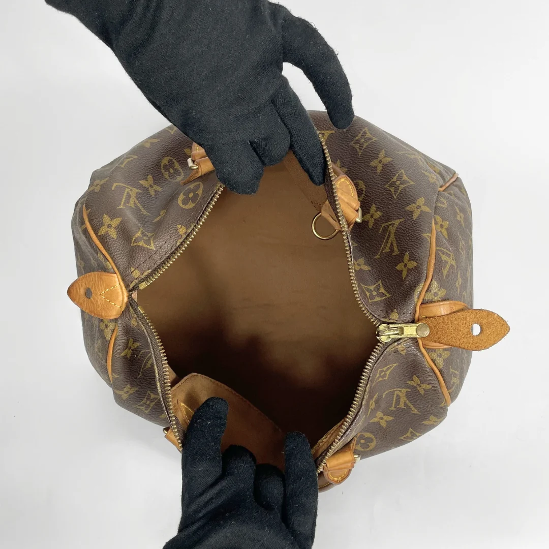 A Louis Vuitton monogram canvas handbag shown open from above, with black gloves holding the zipper area to display the interior. The bag features the classic brown and tan monogram pattern with gold-tone hardware.