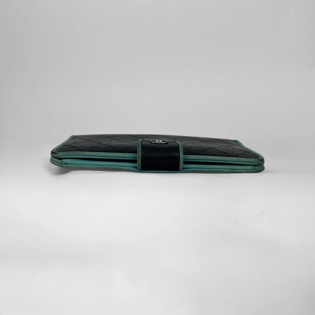 A vintage Chanel long wallet in black quilted leather with a teal green trim and a central black velvet strap featuring a silver-tone interlocking CC logo, set against a plain light gray background.
