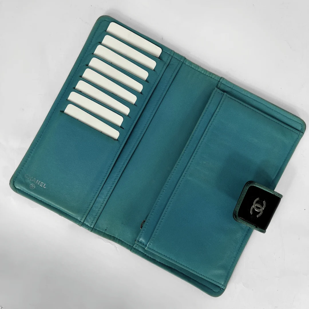 A teal Chanel wallet shown open, featuring multiple white card slots on one side, a large card pocket on the other, and a black leather CC turn-lock closure on the front.