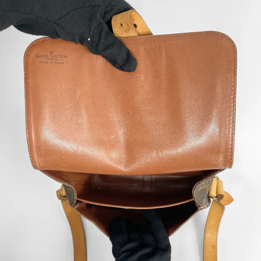 A Louis Vuitton tan leather shoulder bag shown open, with black gloved hands holding it against a plain white background. The Louis Vuitton logo is debossed on the upper left of the flap.