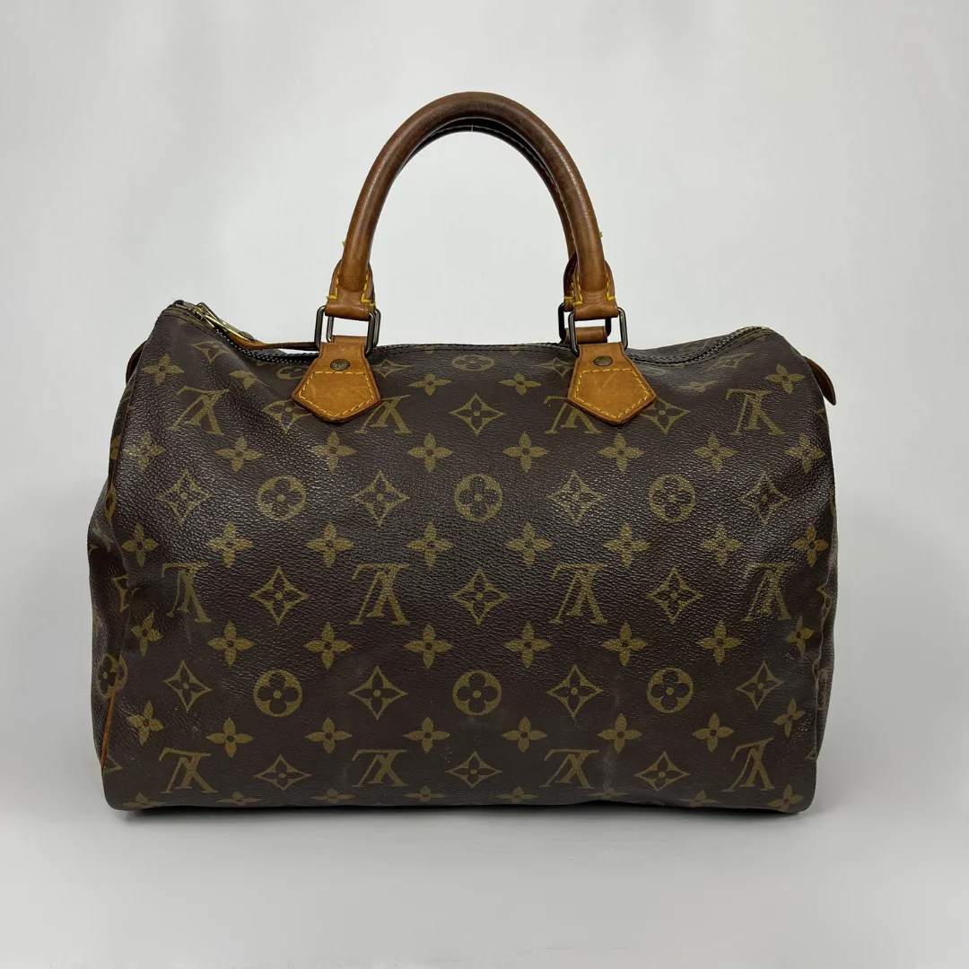 A Louis Vuitton monogram canvas speedy bag with brown leather handles and gold-tone hardware, photographed against a neutral light gray background.