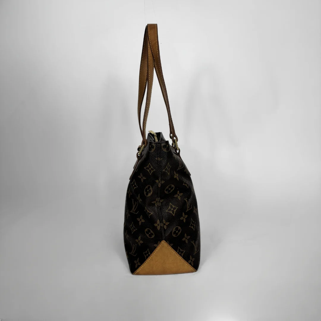 A Louis Vuitton monogram canvas bucket bag with brown leather trim and shoulder strap, standing upright against a light gray background.