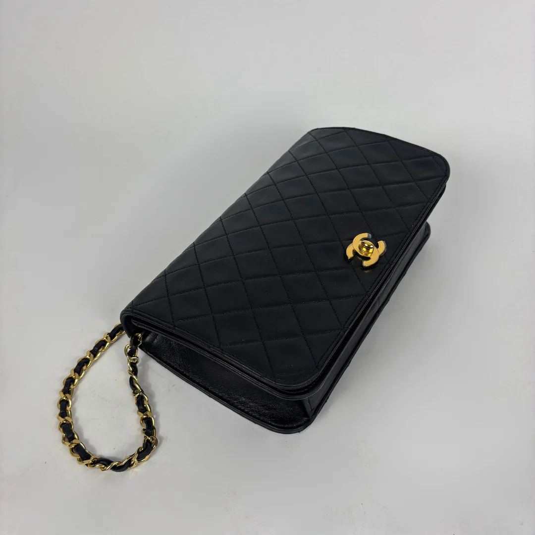 A black quilted leather clutch bag with a gold-tone interlocking CC logo and a gold-and-black chain strap, photographed on a light gray background.