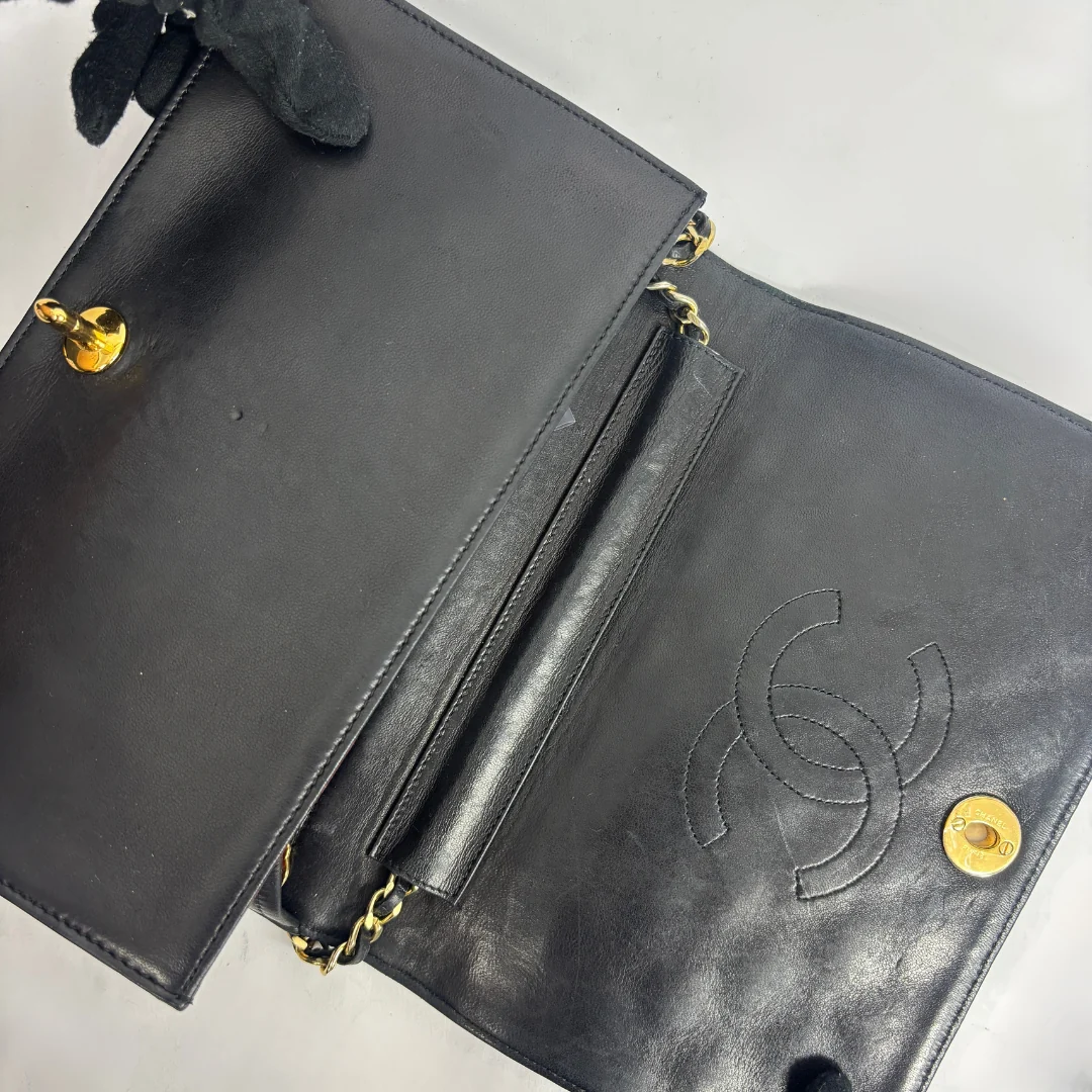 A black leather clutch bag with gold-tone hardware, shown open to reveal the interior. The right side features an embossed interlocking CC logo, characteristic of Chanel.