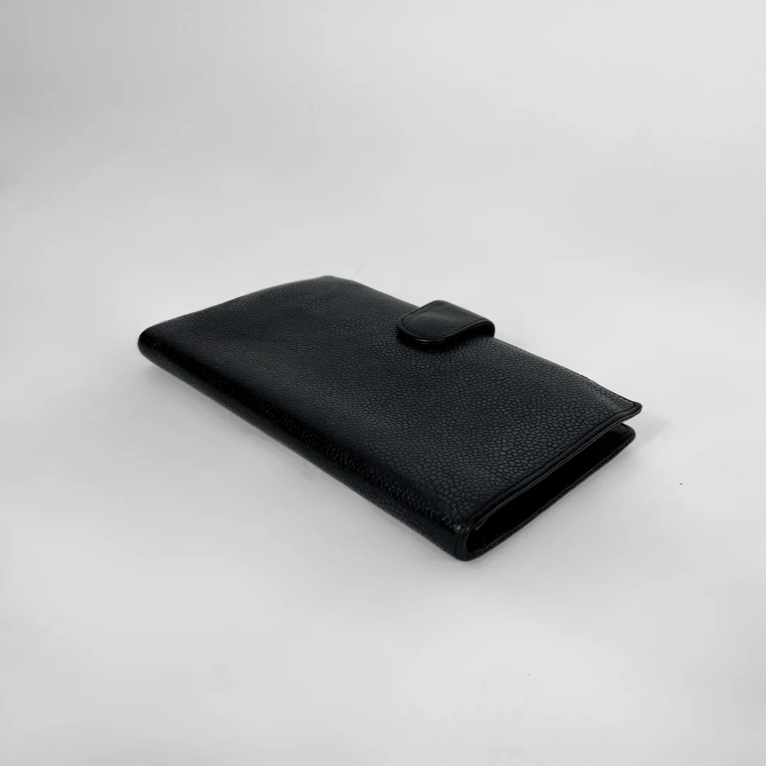 A black textured leather wallet with a small tab closure on the front, photographed on a light gray studio background.