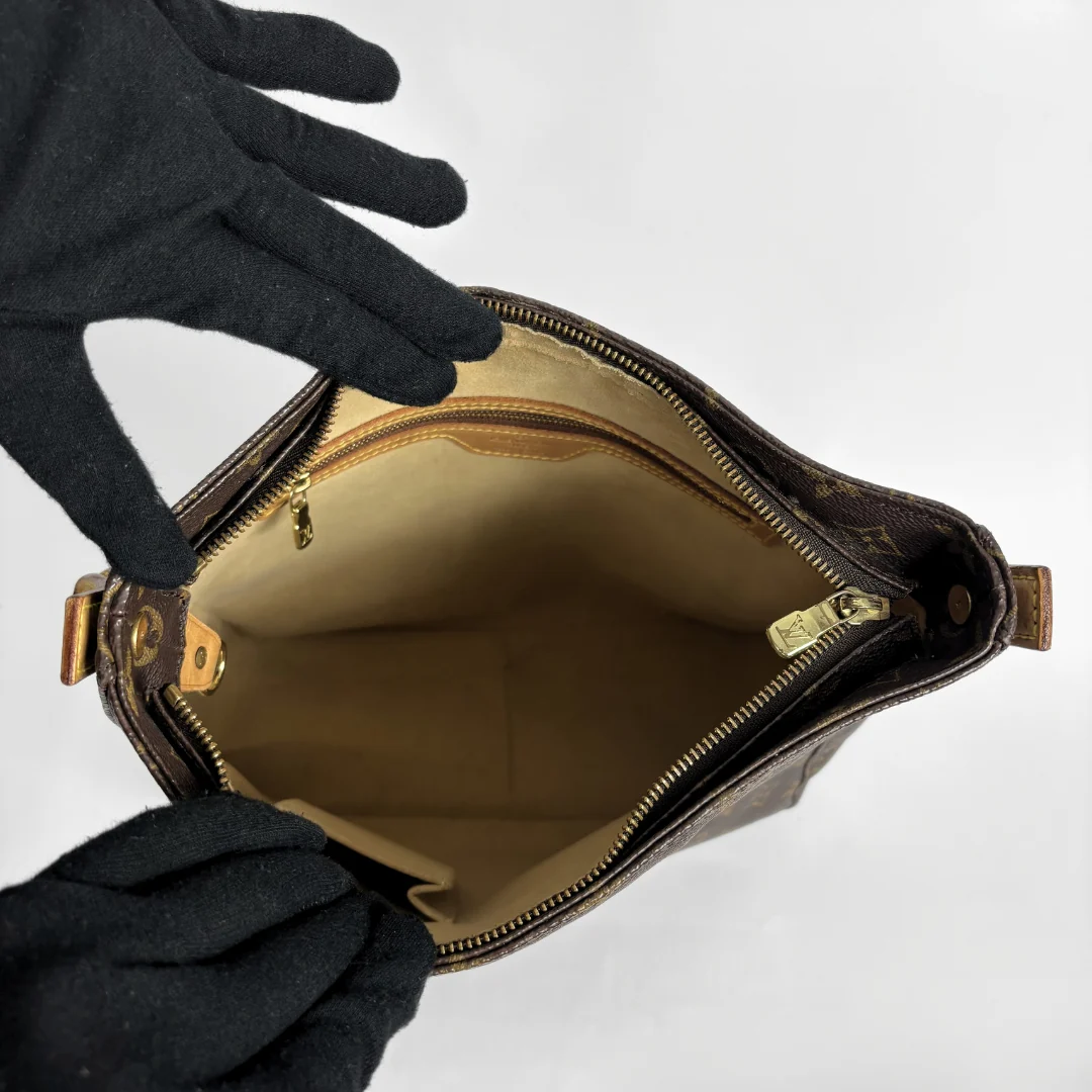 A brown monogram handbag shown open from above, with a beige interior lining and gold-tone zipper hardware. Two black gloves are holding the bag open against a plain light background.