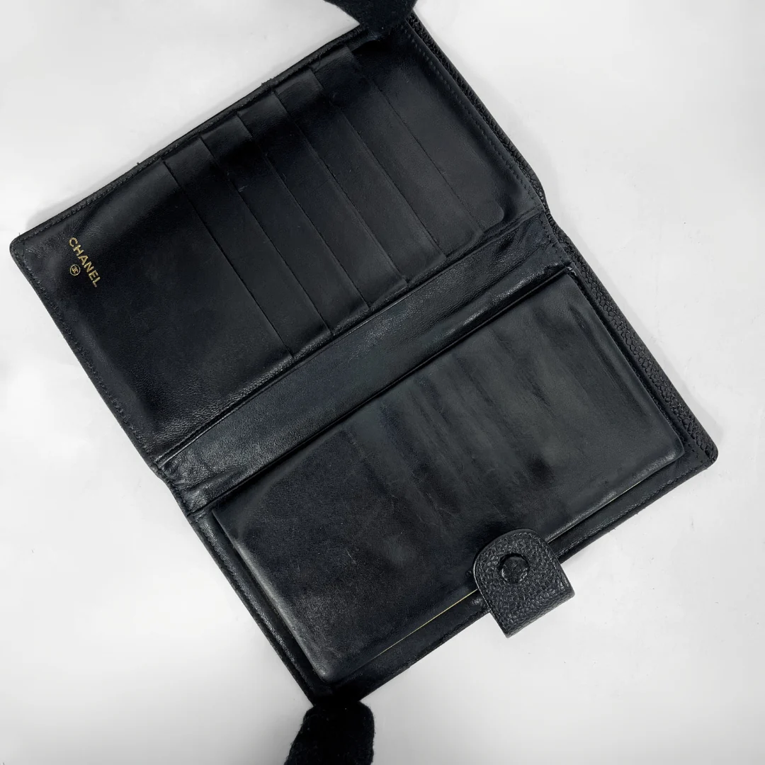 An open black leather wallet with multiple card slots and a large slip pocket, featuring a small gold CHANEL logo on the left side, photographed on a white background.