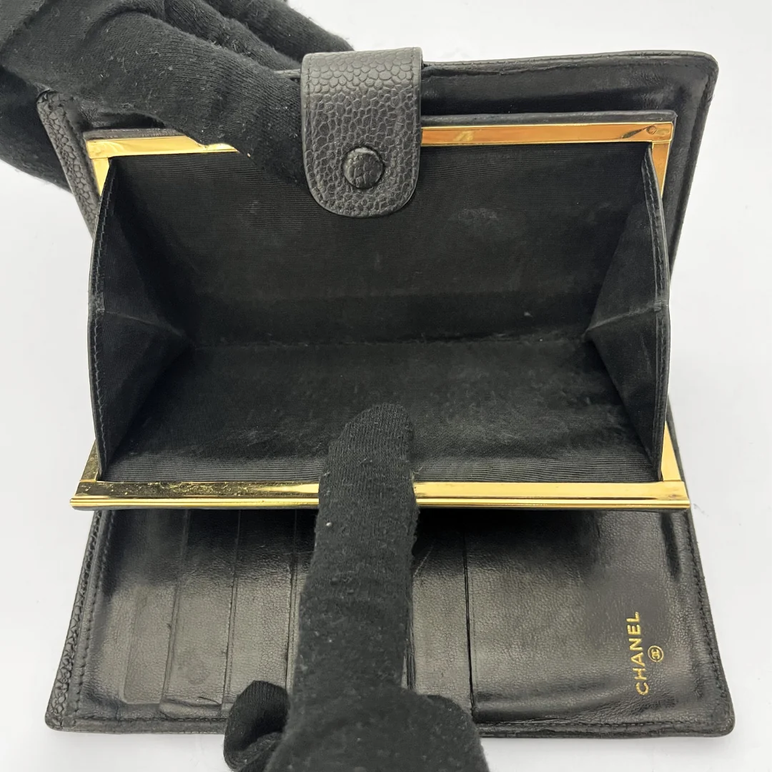 A black leather wallet with gold-tone metal frame details, shown open with gloved hands pointing inside to display the card slots and interior. The Chanel logo is visible in gold on the lower right interior.