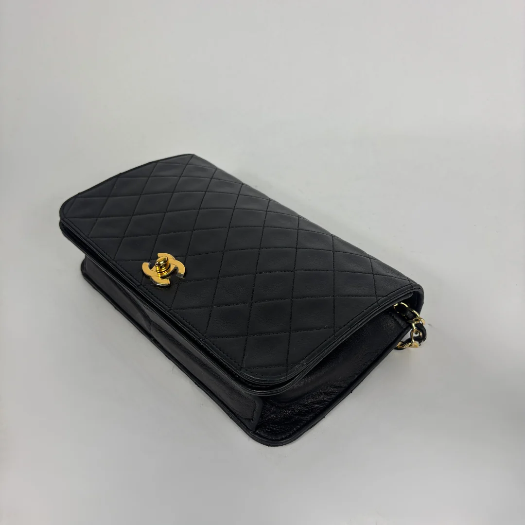 A black quilted leather shoulder bag with a gold-tone interlocking CC clasp, characteristic of Chanel, shown on a neutral light gray background.